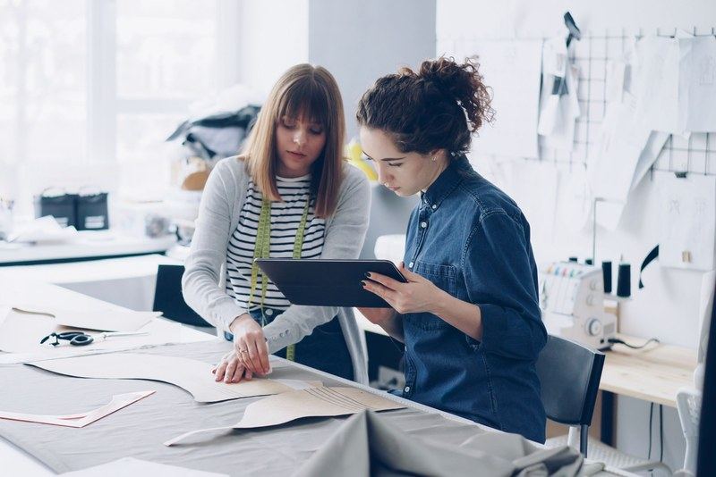 Two fashion designers reviewing a tablet over pattern pieces in a bright design studio